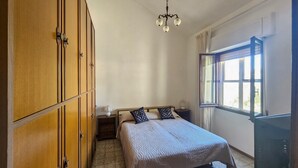 A bedroom with a bed, a wardrobe, a nightstand, a lamp, a window with curtains, and a chandelier. - Casa Bianca Villaputzu (Villaputzu)
