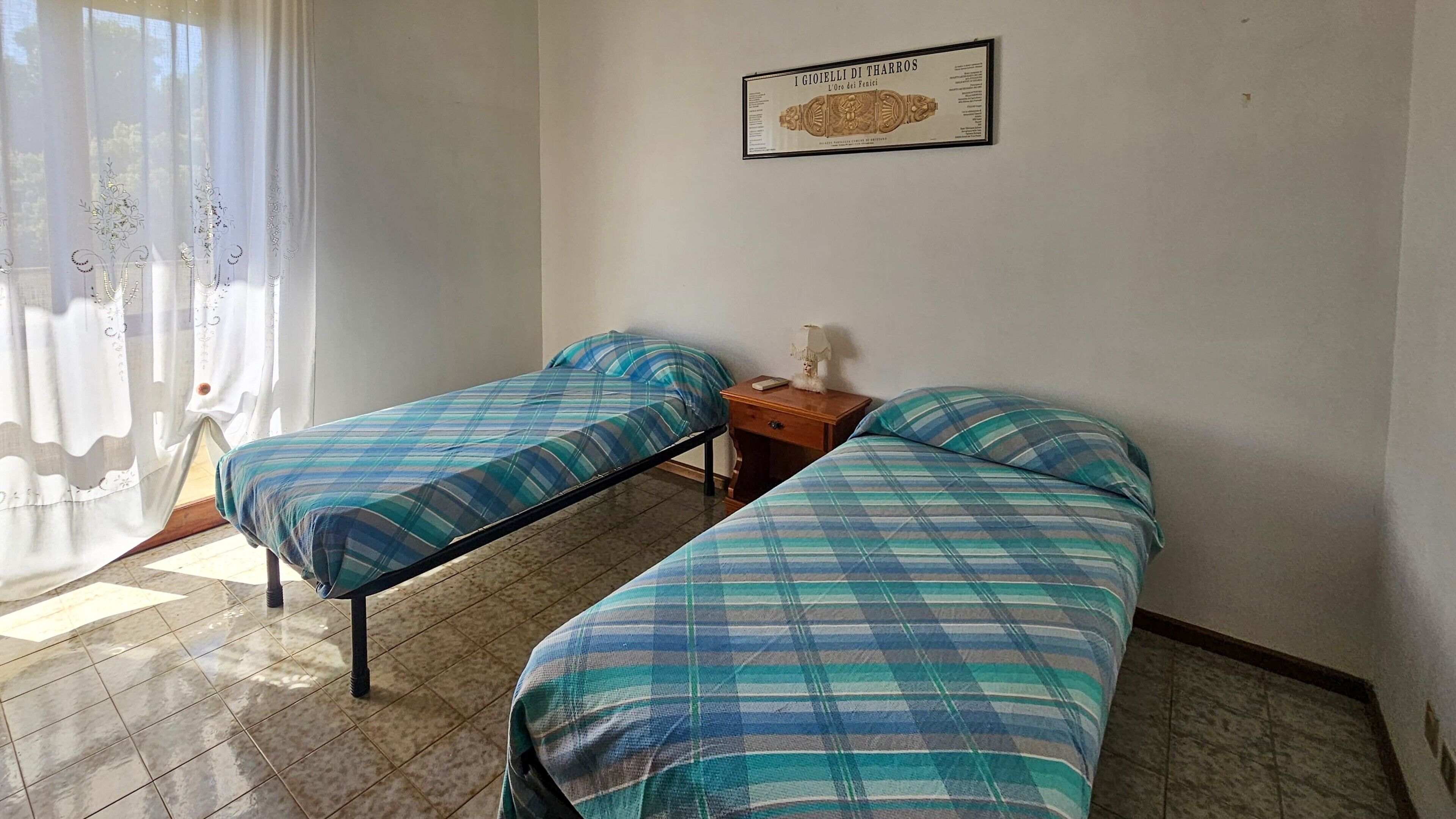 A room with two beds, a wooden nightstand, a framed certificate on the wall, and a window with sheer curtains.