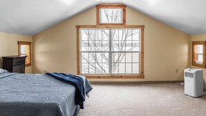 Un dormitorio con techo inclinado, un ventanal con vistas a árboles desnudados, una cama con manta azul y un calentador portátil.