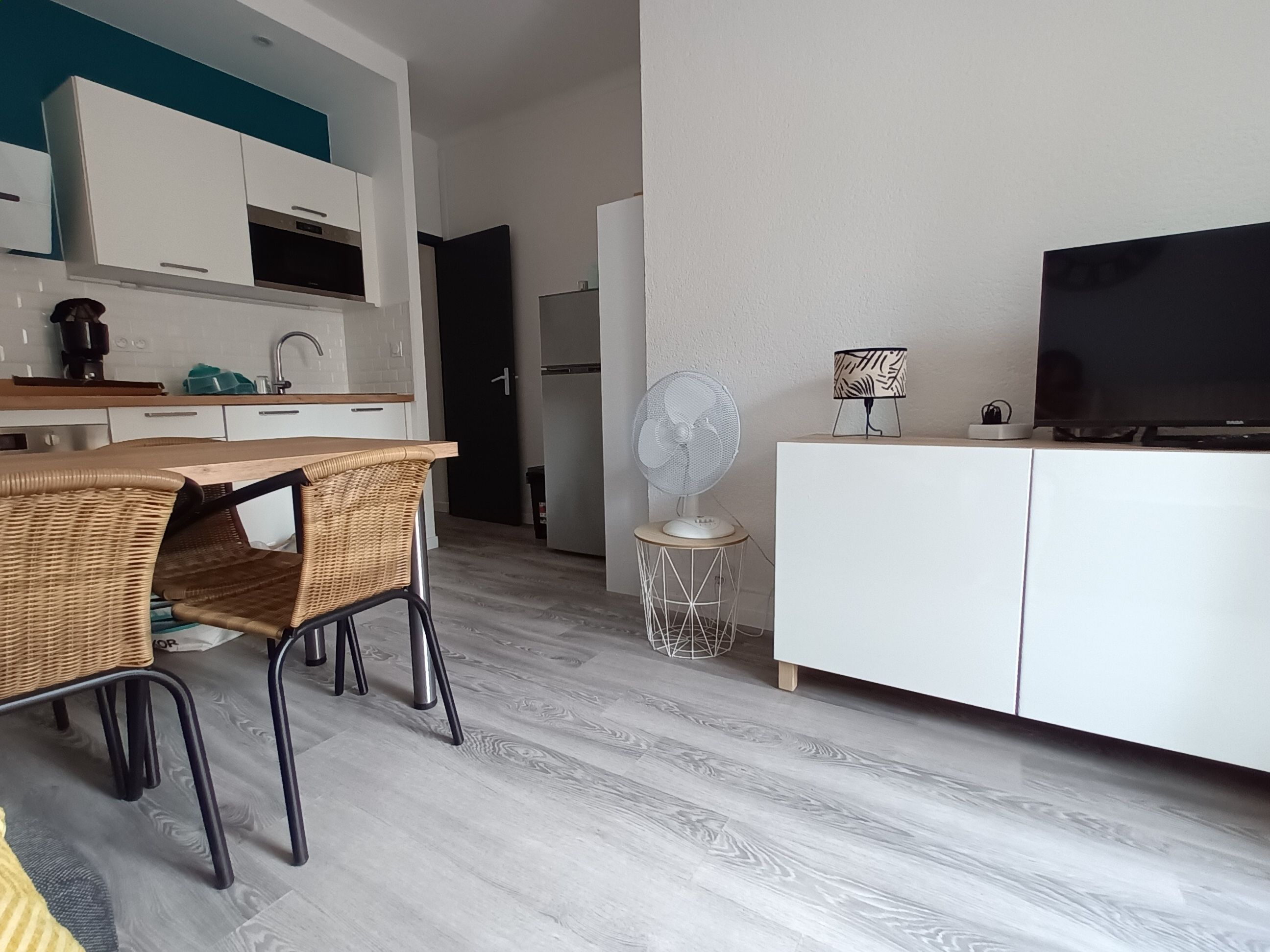 Una cocina moderna con gabinetes blancos, una zona de comedor con sillas de mimbre, un ventilador de pie y un televisor de pantalla plana sobre un mueble blanco.