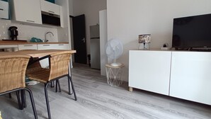 Une cuisine moderne avec des meubles blancs, un coin repas avec des chaises en osier, un ventilateur sur pied et un téléviseur à écran plat posé sur un meuble blanc.