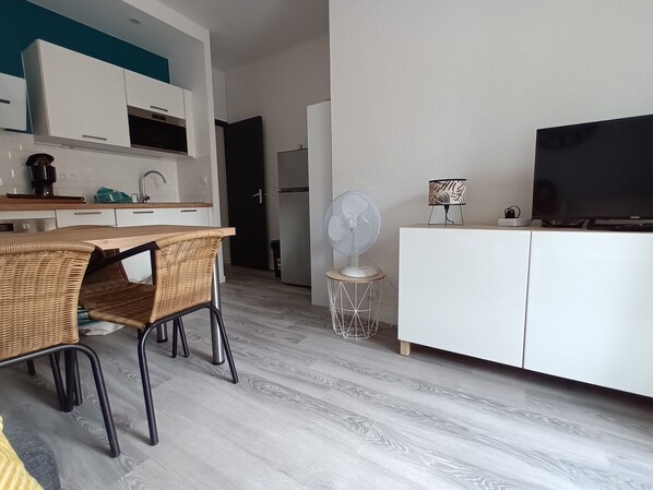 A modern kitchen with white cabinets, a dining area with wicker chairs, a standing fan, and a flat-screen TV on a white cabinet.