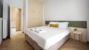 A neatly made bed with a white comforter and pillows, a bedside table with a plant, and a wardrobe with a wooden door. - Spacious & Well Designed•2BR Apartment in Pangrati (Athina)