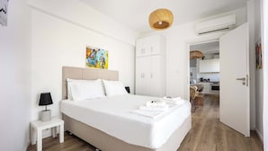 A modern hotel room with a large bed, a bedside table with a lamp, a wardrobe, and a kitchen visible through an open door. - Bright Stylish 2BR | Acropolis View Balcony | King Bed (Athina)
