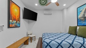 A bedroom with a bed, a bench, a chair, a ceiling fan, a TV, and artwork on the walls. - New 1 BR Condo - Rooftop Pool - Oceanview (Puerto Vallarta)