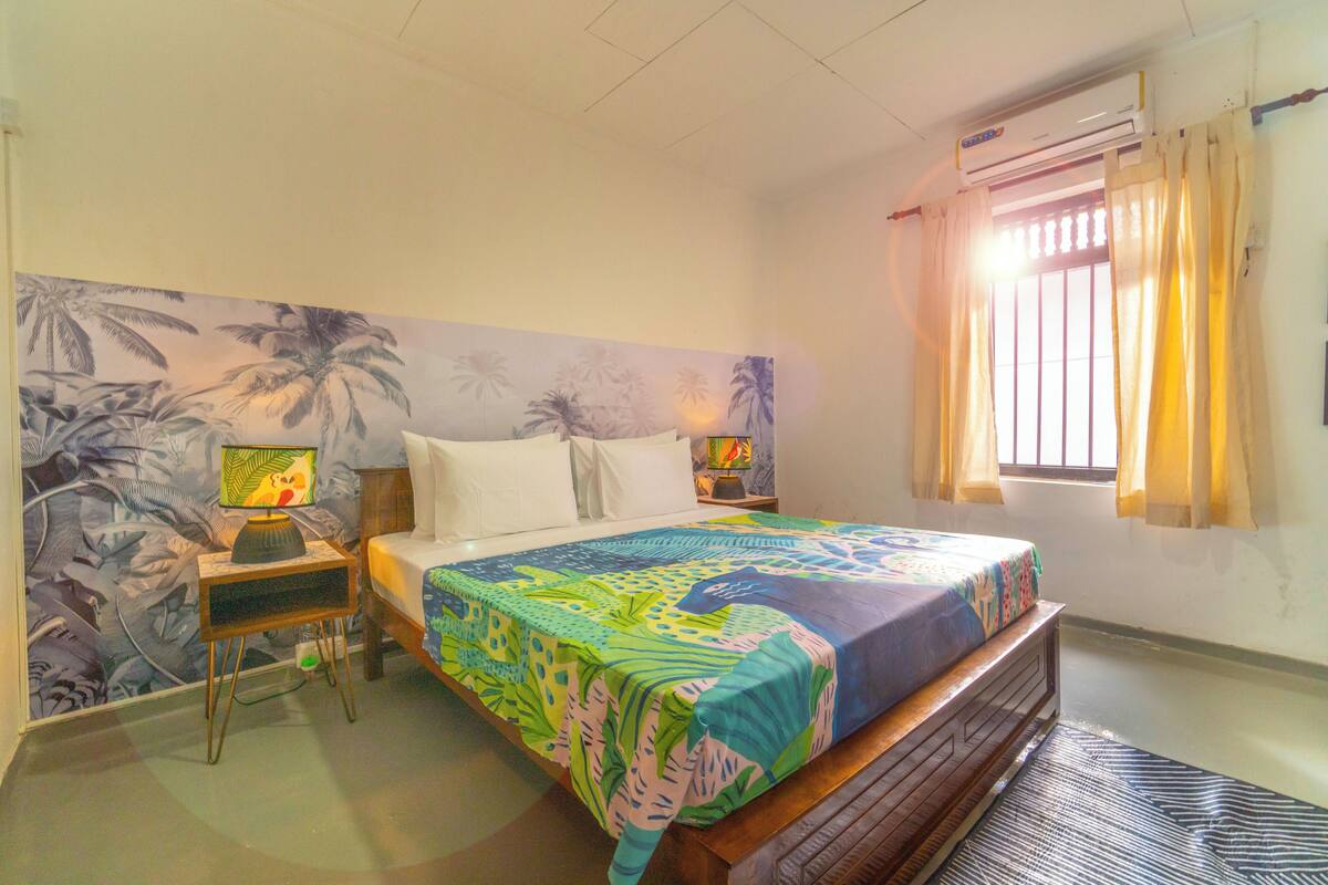 A bedroom with a bed, a nightstand, a lamp, a window with curtains, and a wall with a tropical mural.