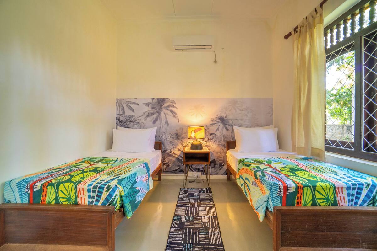 A room with two beds, a nightstand with a lamp, a patterned rug, and a window with curtains.