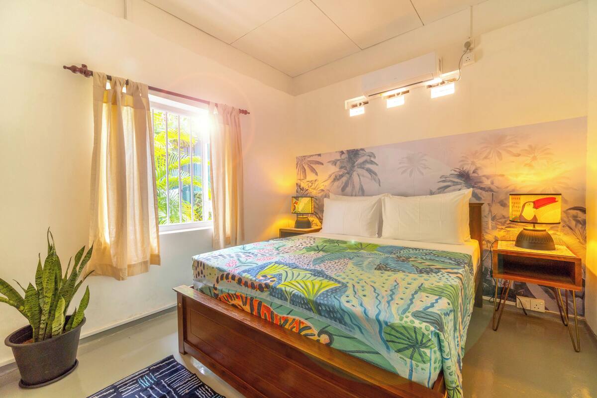 A bedroom with a bed, a nightstand, a lamp, a potted plant, and a window with curtains.