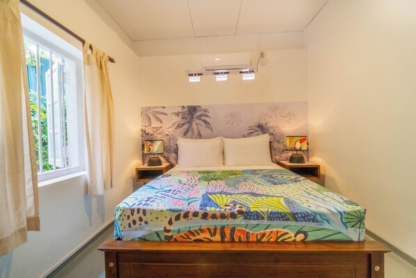 Una camera da letto con un letto, due comodini con lampade, una finestra con tende e una parete con un murale tropicale.