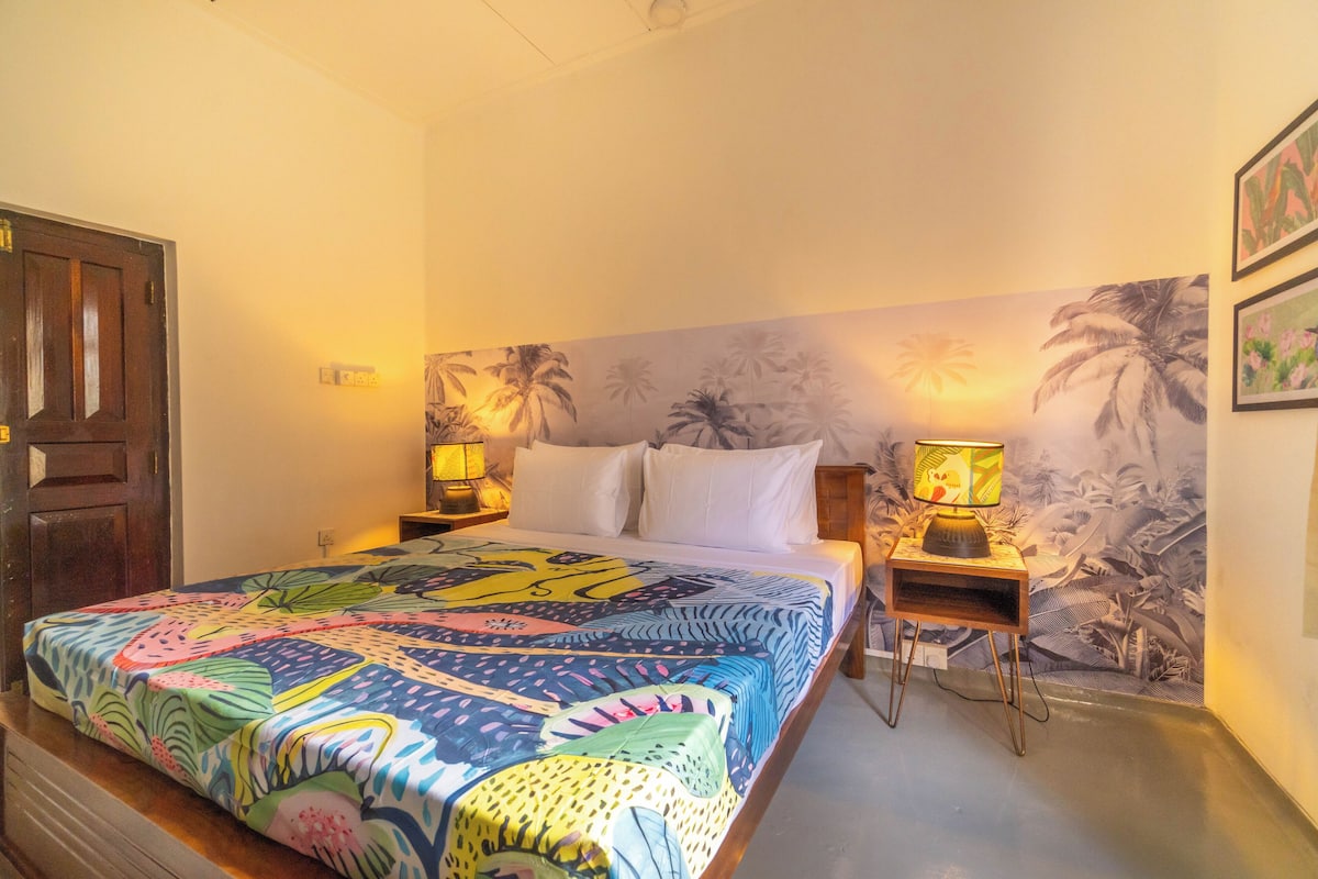 A bedroom with a bed, two bedside tables with lamps, a door, and a wall with a tropical mural.