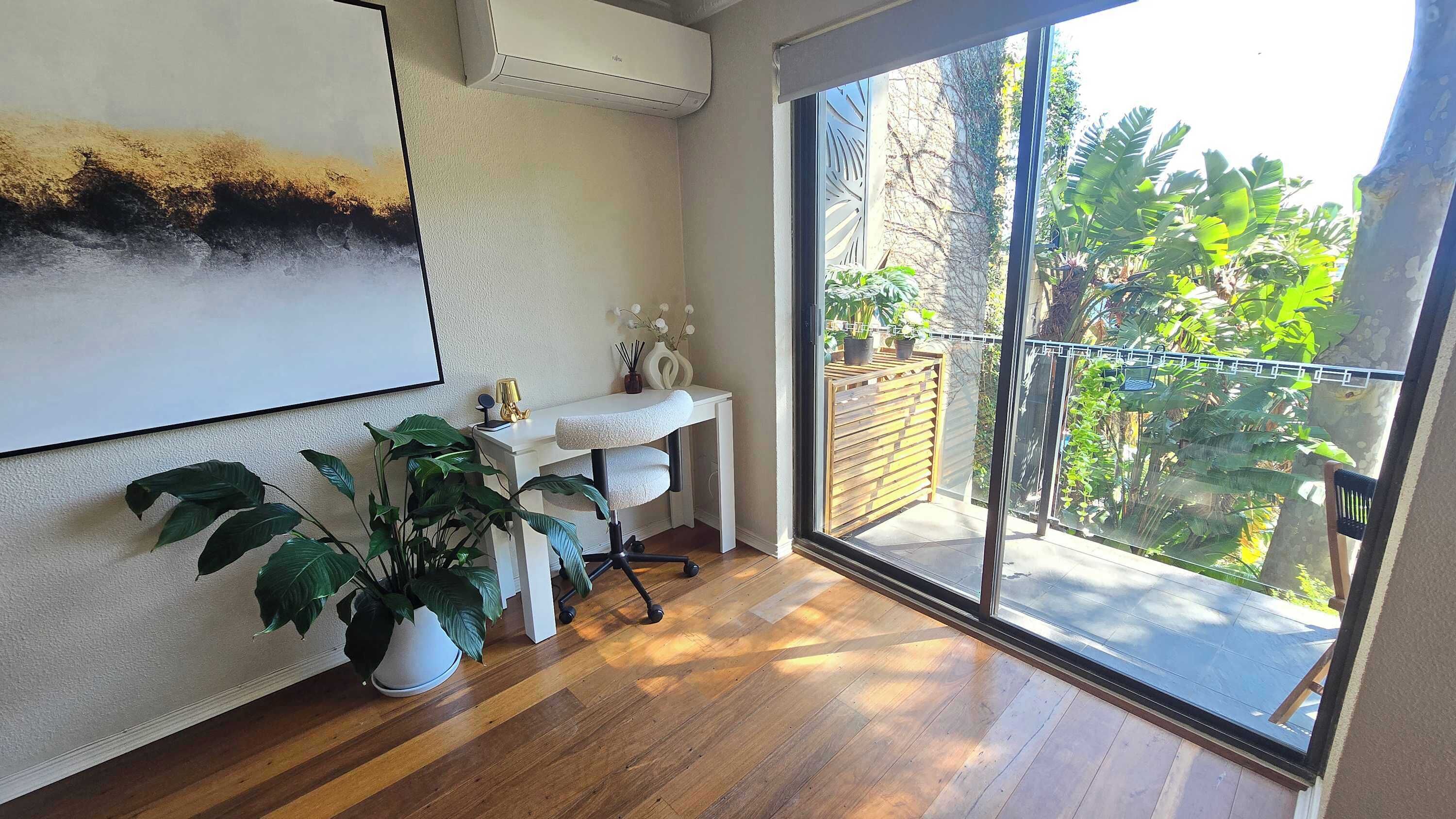 A room with a wooden floor, a white desk, a potted plant, and a large window with a view of greenery.