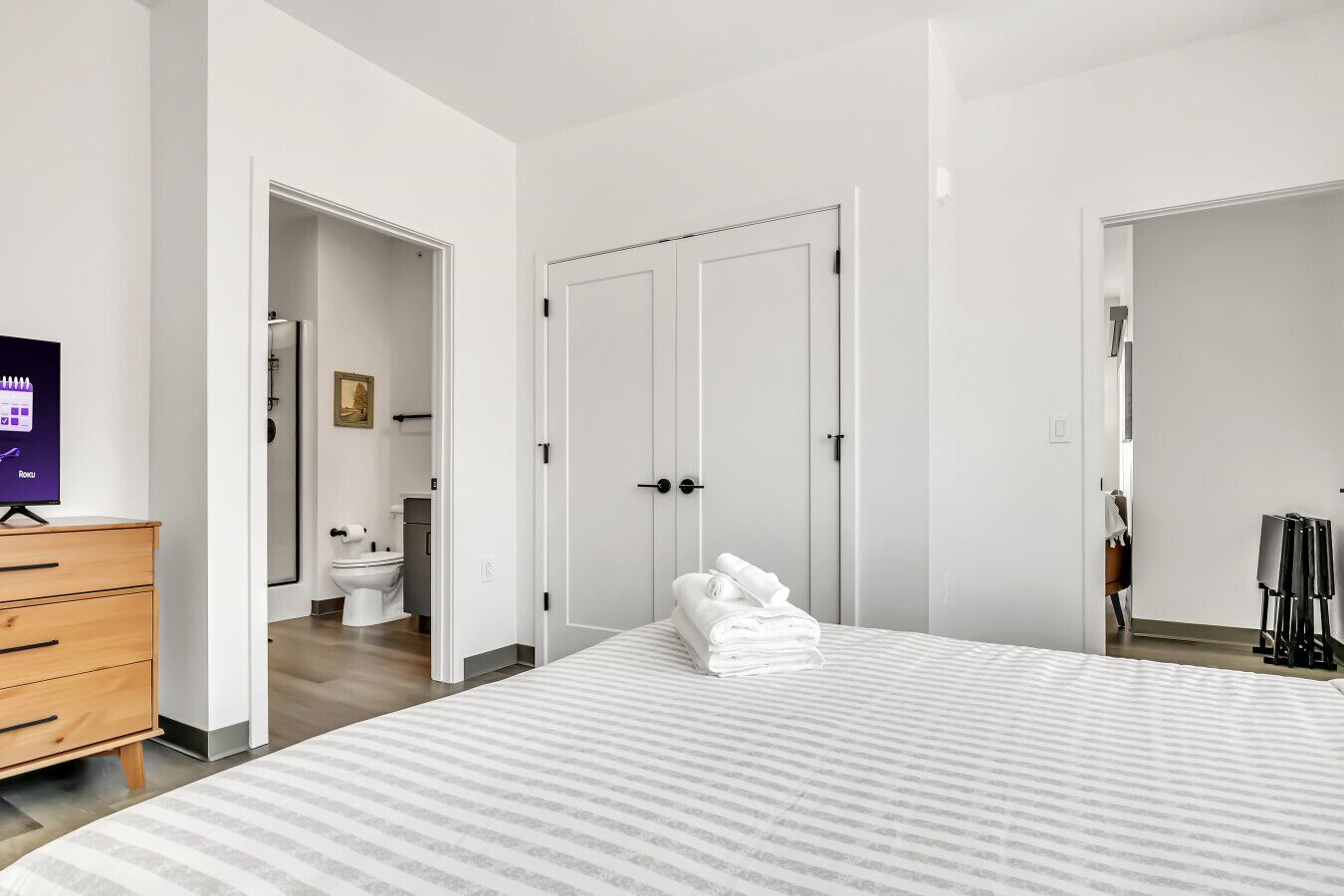 A modern bedroom with a large bed, a wooden dresser, a television, and a bathroom visible through an open door.