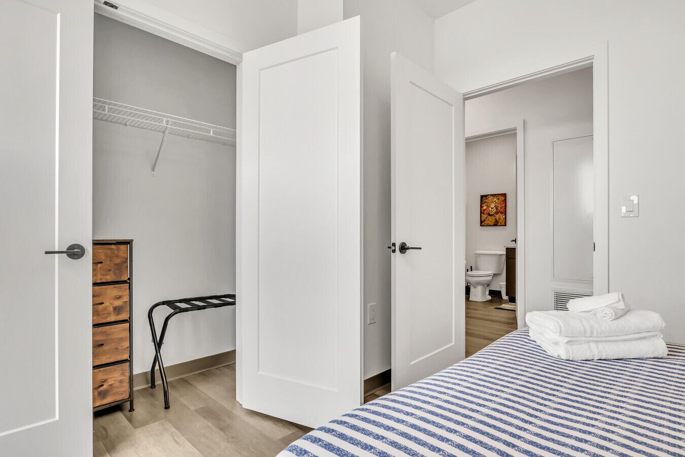 A bedroom with a bed, a wooden dresser, a hanging rack, and a bathroom visible through an open door.