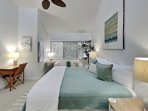 A bedroom with a large bed, two bedside tables with lamps, a desk with a chair, and a window with blinds.