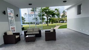 A covered outdoor seating area with wicker furniture and a tiled floor, overlooking a grassy area with trees and buildings. -  White Sand Beach Retreat (Punta Cana)