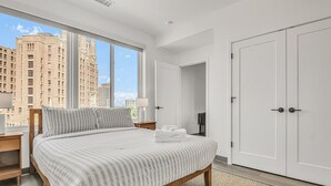 Une chambre d’hôtel moderne avec un grand lit, une vue sur de hauts immeubles, une armoire et une table de chevet avec une lampe.