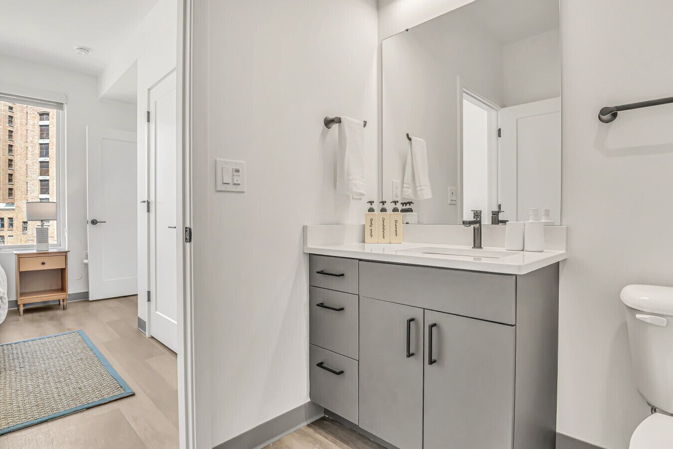 A modern bathroom with a large mirror, white vanity, and a toilet. There are three bottles of toiletries on the vanity.