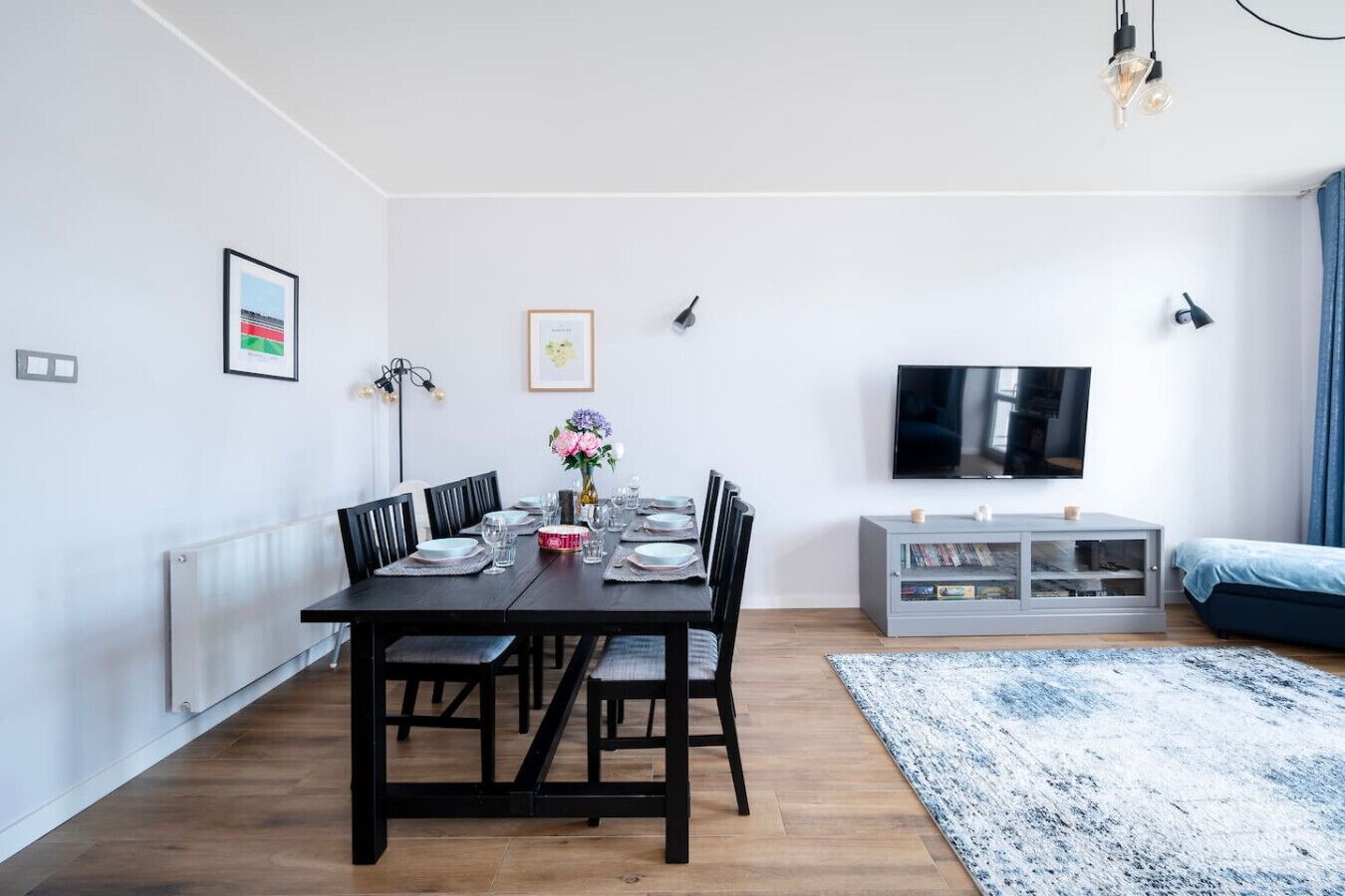 A modern living room with a dining area, a flat-screen TV mounted on the wall, and a grey cabinet with books.