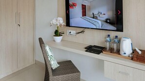 Ein modernes Hotelzimmer mit einem großen Flachbildfernseher an der Wand, einem Korbstuhl, einem kleinen Tisch mit Wasserkocher und einem Schrank darunter.