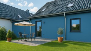 Une maison moderne à l’extérieur bleu, avec un patio comprenant une table et des chaises, ainsi que deux plantes en pot.