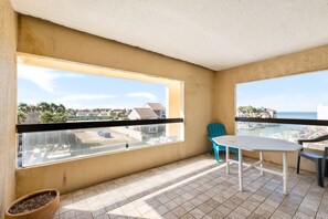 A balcony with a view of the sea and buildings, a white table, two chairs, and a potted plant.