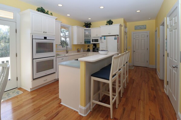 A kitchen with white cabinets, a double oven, a microwave, and a refrigerator. There is a small island with a bowl and a chair.