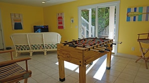 A room with a foosball table, a wicker sofa, a television, and a window with a view of trees.