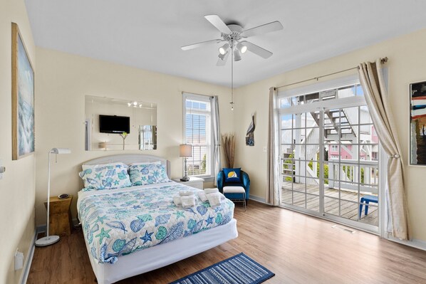 Une chambre à coucher avec un lit, un ventilateur de plafond, une télévision, une fenêtre et une vue sur un balcon.