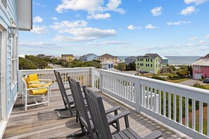 A wooden deck with outdoor furniture, a white railing, and a view of houses and the ocean.
