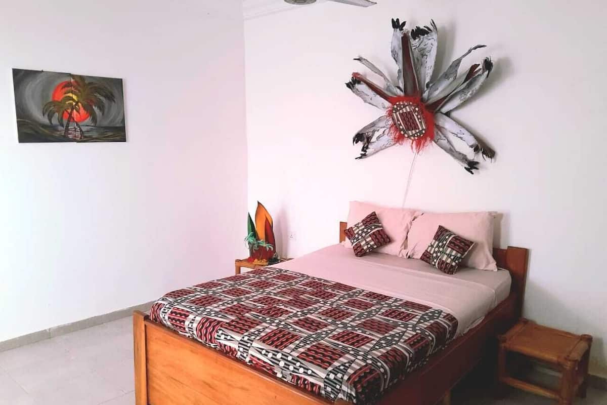 A bedroom with a wooden bed, a patterned bedspread, a wall-mounted decorative piece, and a painting on the wall.