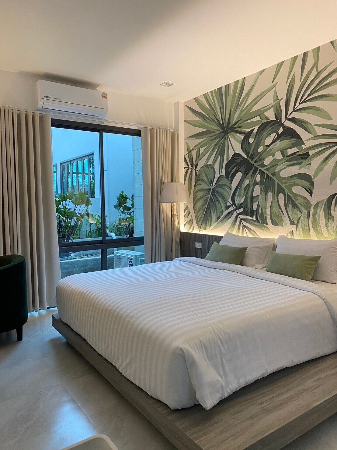 A hotel room with a large bed, a view of the outdoors, and a tropical leaf mural on the wall.