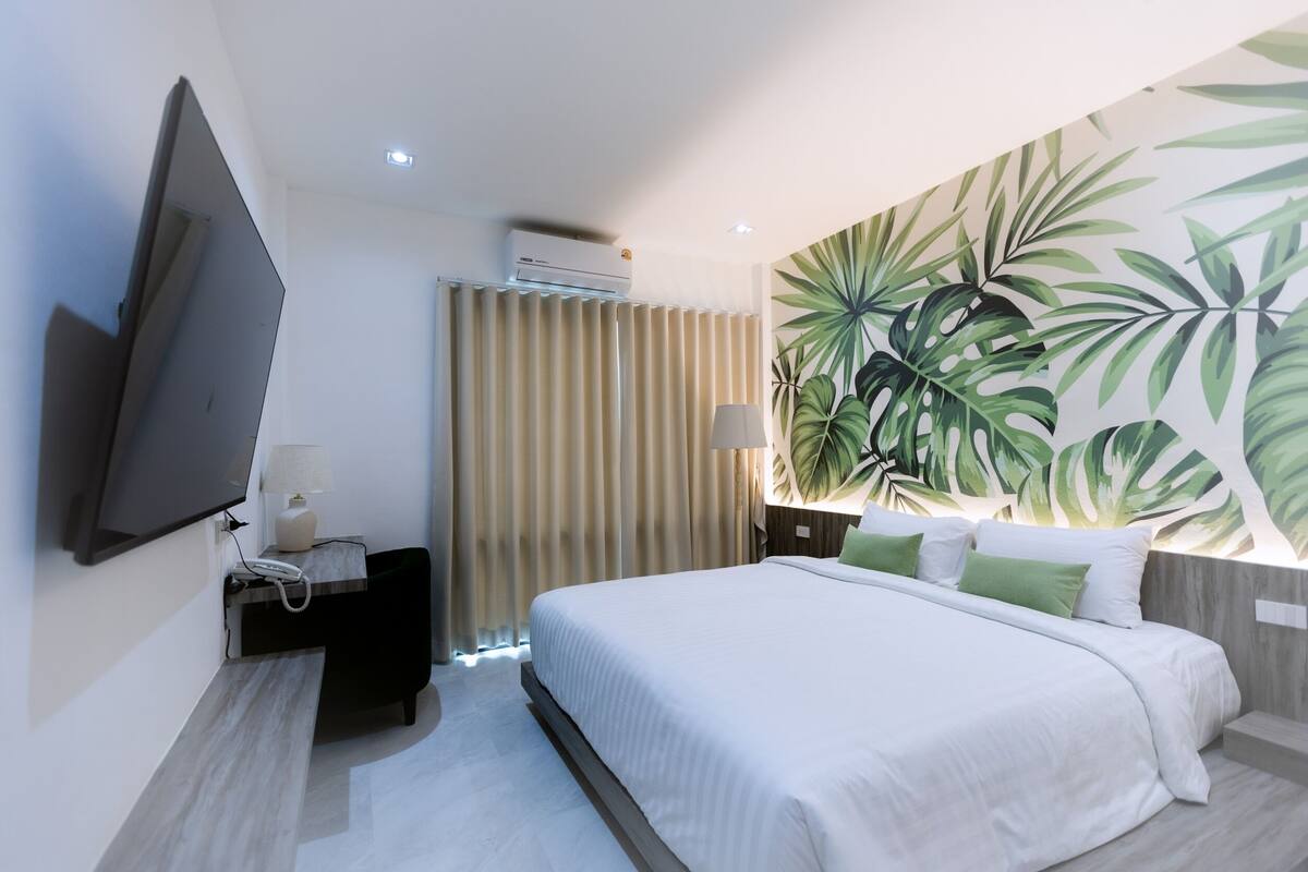 Ein modernes Hotelzimmer mit einem großen Bett, einem flachbildfernseher an der Wand, einem Nachttisch mit Lampe und einem Wandbild mit tropischem Blattmuster.