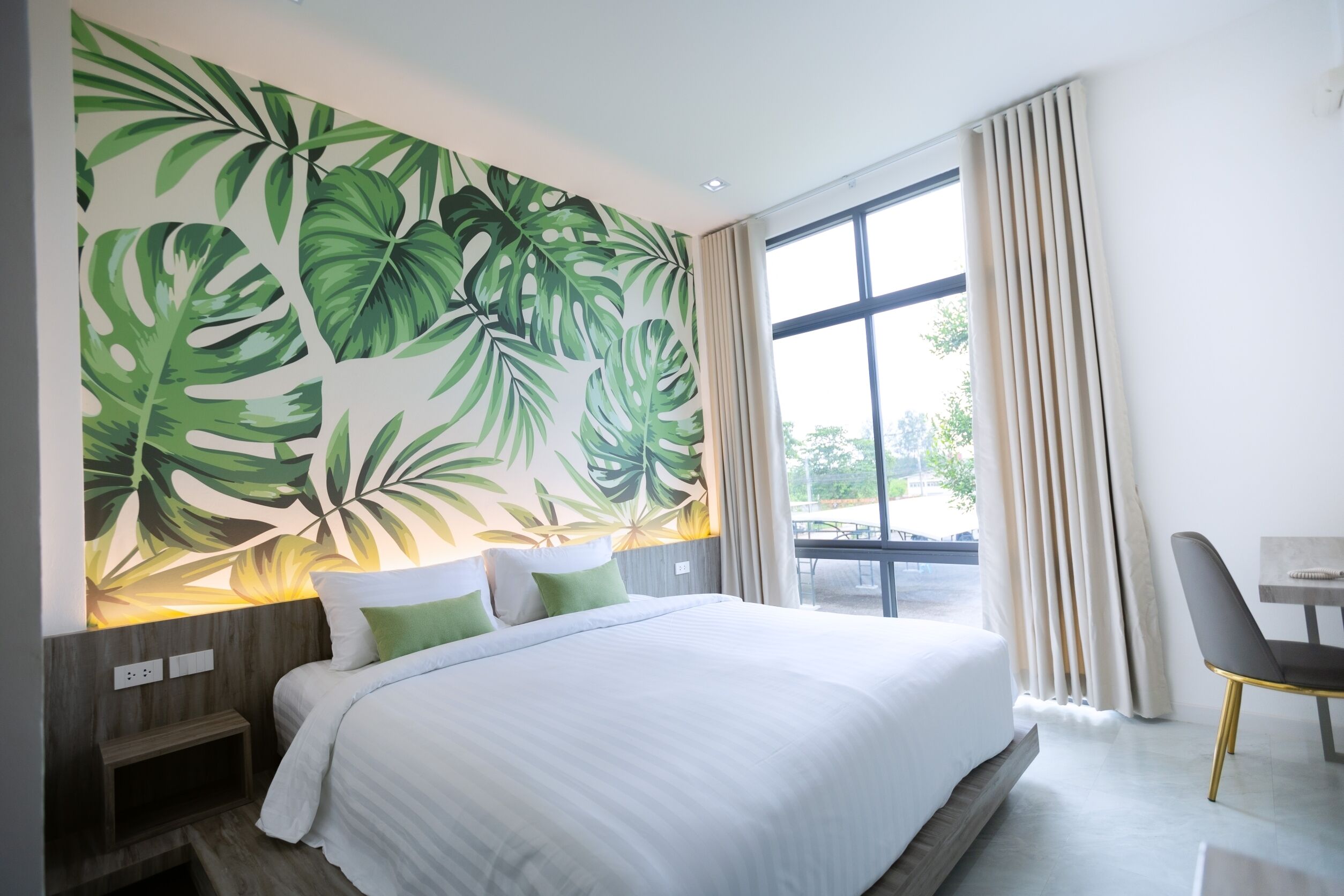 Une chambre d’hôtel avec un grand lit, une vue sur l’extérieur par une fenêtre et un mur décoré d’un motif de feuilles tropicales.