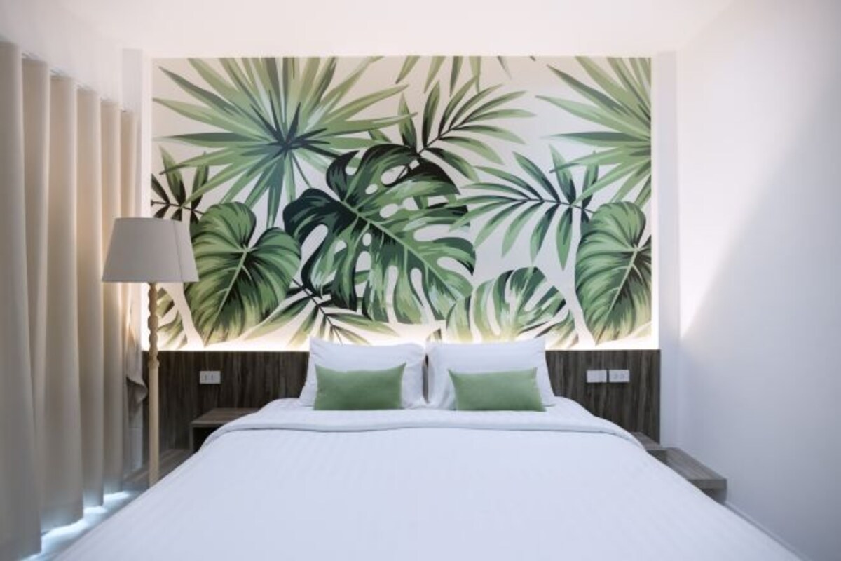 Une chambre d’hôtel avec un grand lit, une table de chevet, une lampe et un mur orné d’un motif de feuilles tropicales.