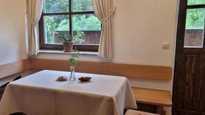 Una habitación con una mesa dispuesta para dos personas, una ventana con cortinas, un banco de madera y una pequeña planta en maceta en el alféizar.