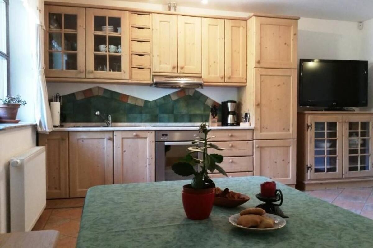 Ett kök med träskåp, kakel ovanför arbetsbänken, en kaffebryggare, en tv och ett matbord med en växt och en tallrik mat.