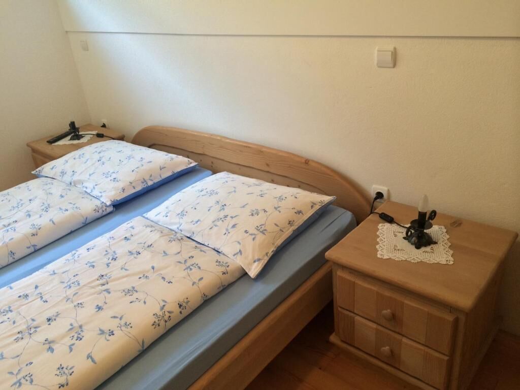 A small, simple room with two single beds, each with floral-patterned bedding. There is a wooden nightstand with a lamp and a small drawer.