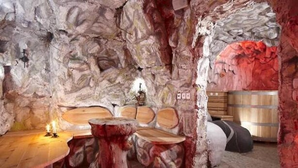 Un interior rústico con pared de piedra, asientos de madera y una vela sobre la mesa.