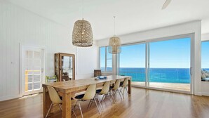 A spacious dining area with a wooden table and chairs, large windows with a view of the ocean, and two hanging wicker lamps. - Fairwinds Manyana Stunning Views (Manyana)
