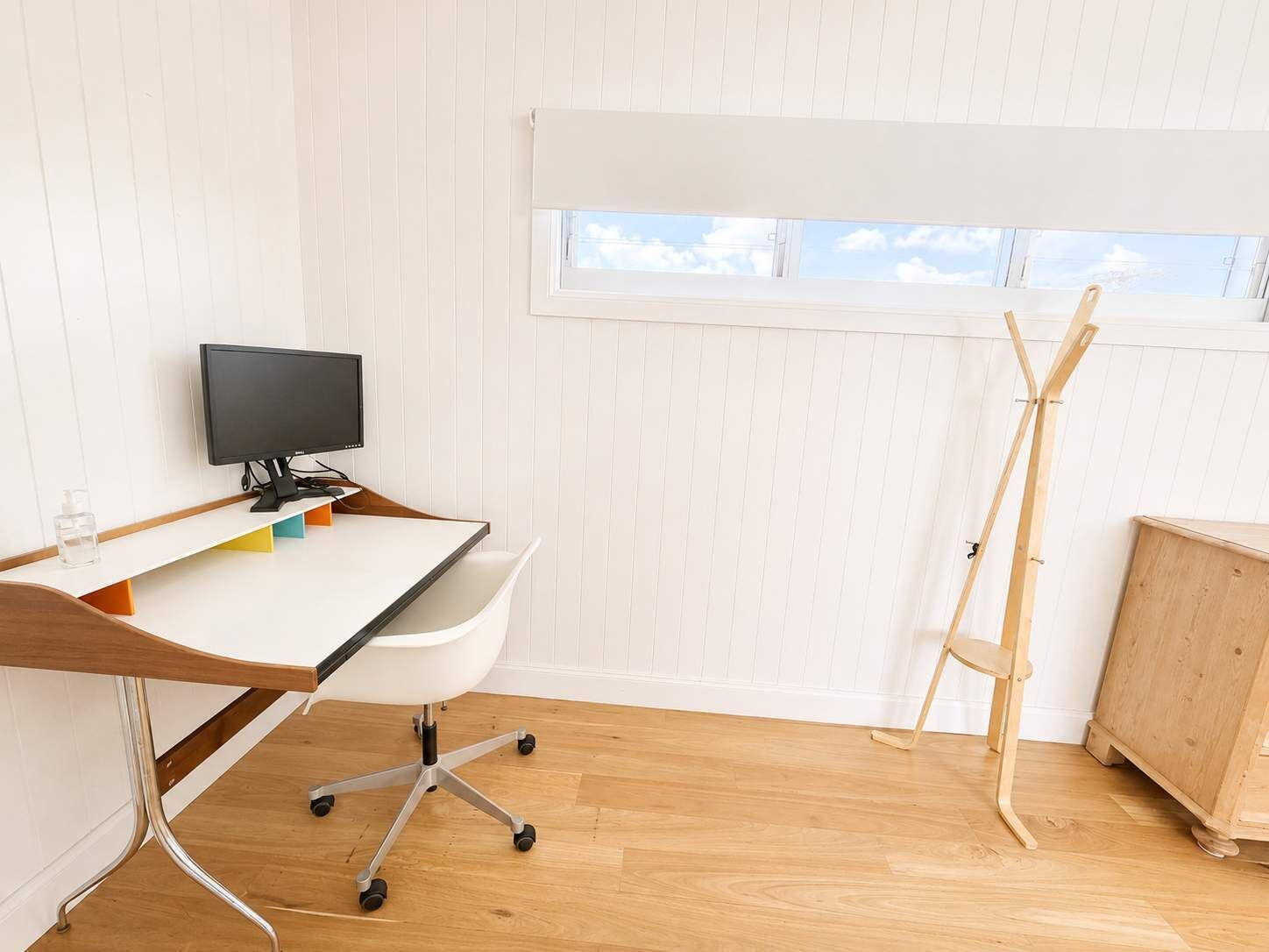 Una habitación con un escritorio blanco, un monitor de computadora, una silla blanca, un caballete de madera y un mueble de madera.