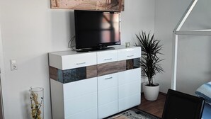 Een woonkamer met een flatscreen televisie op een witte kast, een potplant en een lijst met een foto aan de muur.