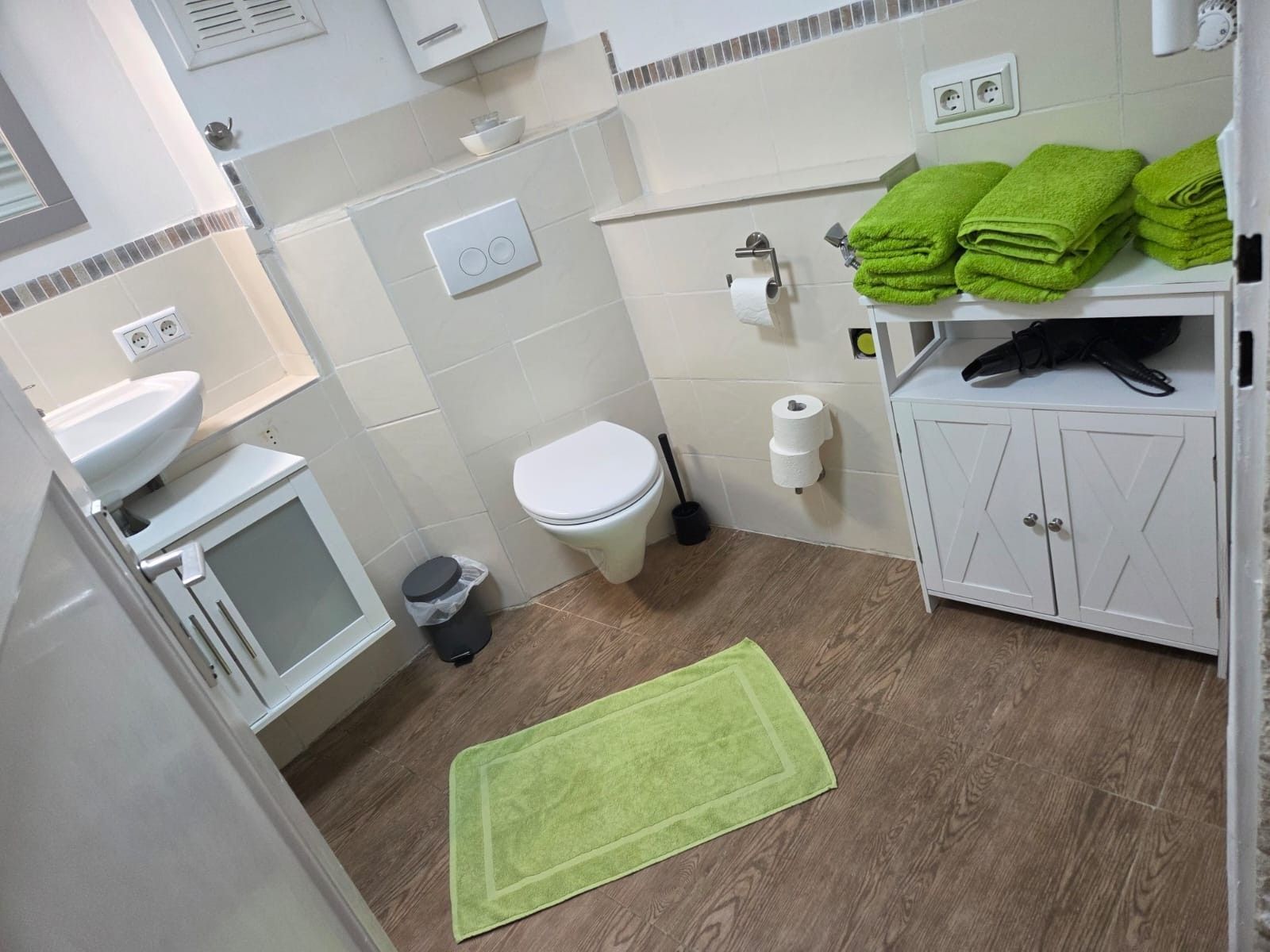 A bathroom with a toilet, sink, and cabinet, a green rug, and a stack of green towels.