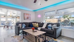 A modern living room with a grey sofa, a coffee table, and a colorful rug. There is a ceiling fan, a window with blinds, and a neon sign on the wall. - Heated Pool | Putting Green | 7 Min Old Town (Scottsdale)