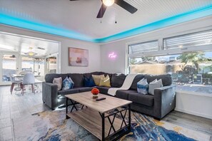 A modern living room with a grey sofa, a coffee table, and a colorful rug. There is a ceiling fan, a window with blinds, and a neon sign on the wall. - Heated Pool | Putting Green | 7 Min Old Town (Scottsdale)