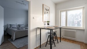 A modern bedroom with a bed, a small table with stools, a radiator, and a window with blinds. - Bright & Cozy 1BR Home near Downtown (Lahti)