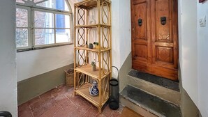 Una stanza con una porta in legno, uno scaffale di vimini con piante, una finestra e un pavimento piastrellato.