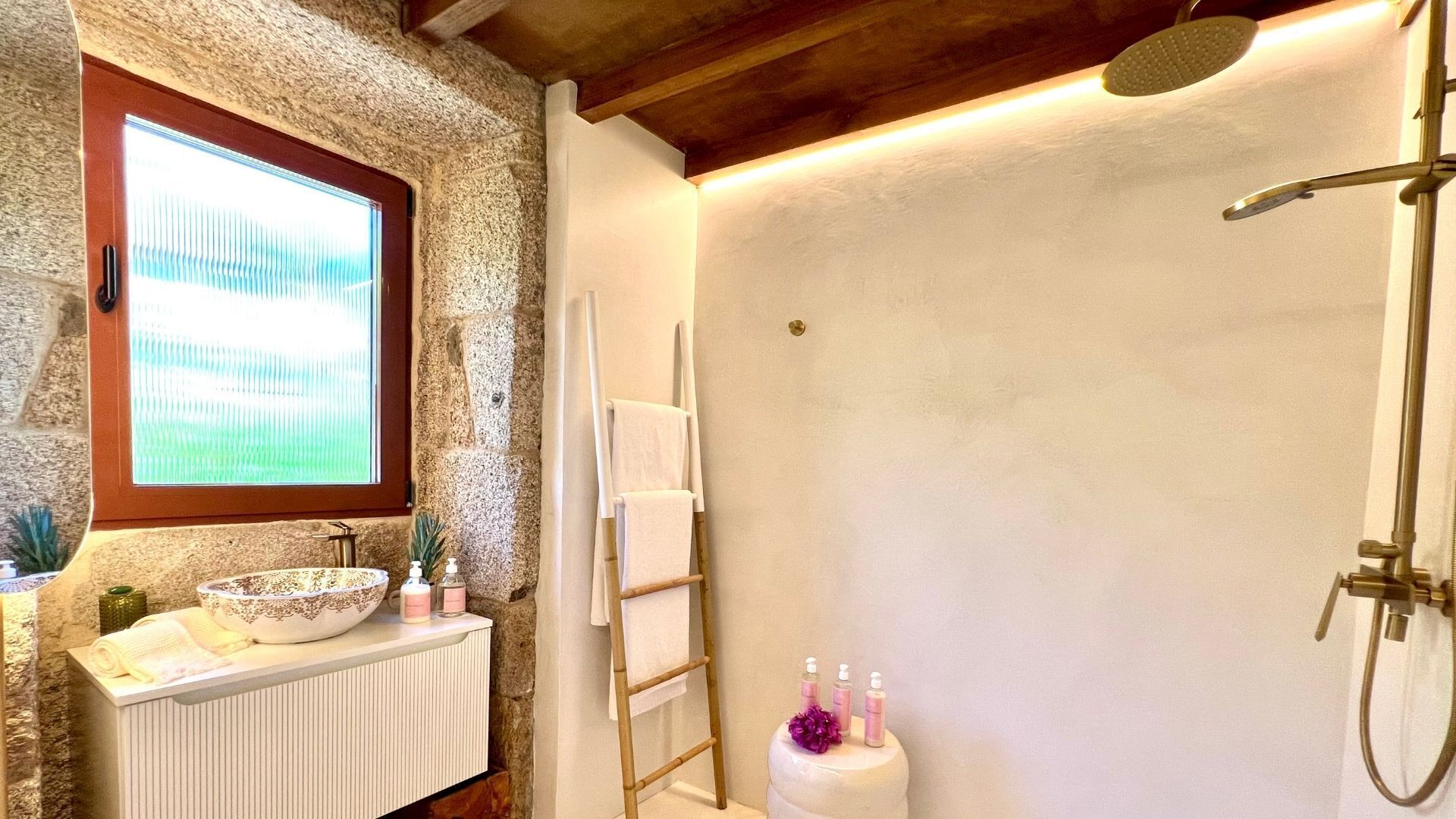 Un baño con pared de piedra, una ventana, una bañera, un perchero para toallas y artículos de aseo sobre el mostrador.