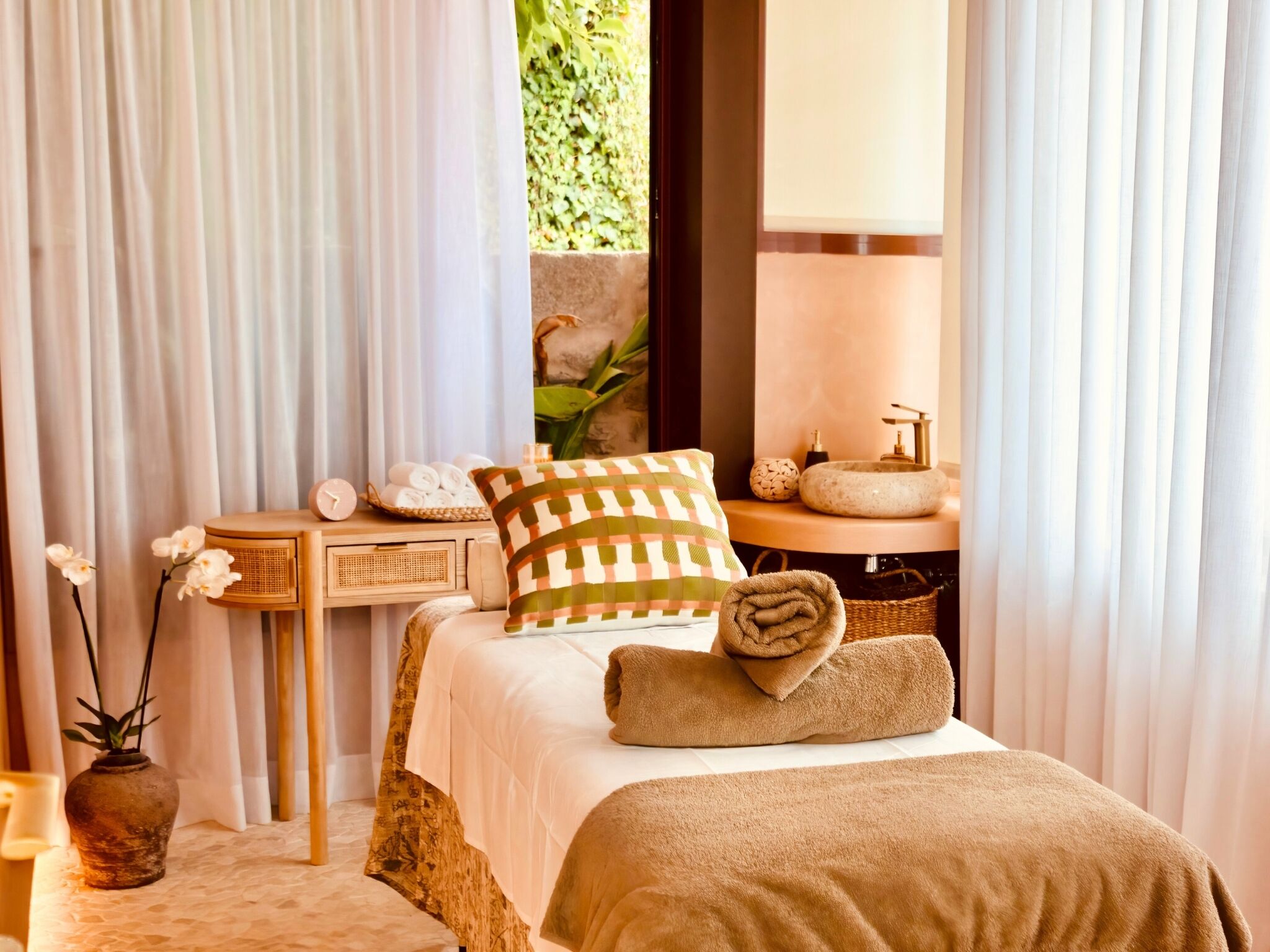 A well-lit bedroom with a bed, a bedside table with a clock, a vase with flowers, and a view of a stone wall and greenery outside.