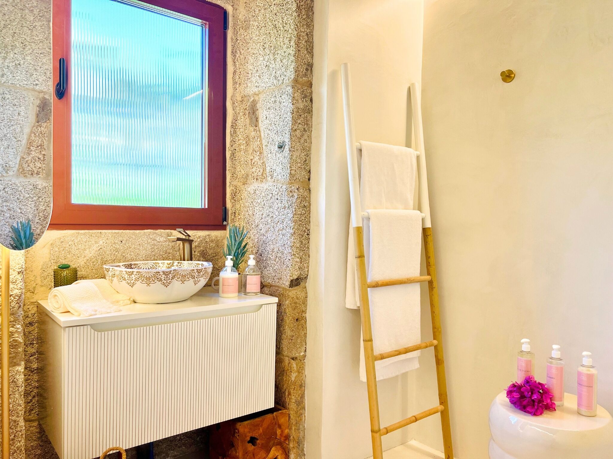 A bathroom with a stone wall, a window with blinds, a white sink with a decorative bowl, a wooden ladder, and toiletries.