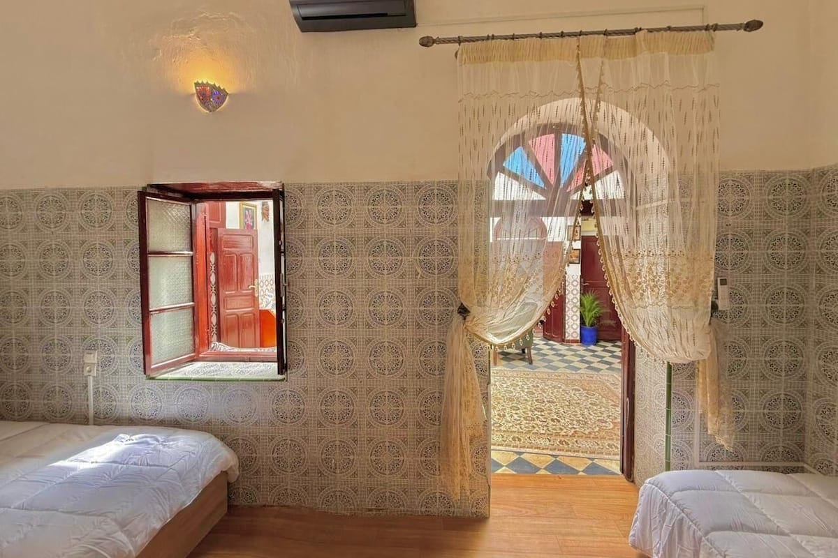 A room with two beds, a window with a view of a colorful outdoor area, a wall-mounted air conditioner, and a decorative light fixture.